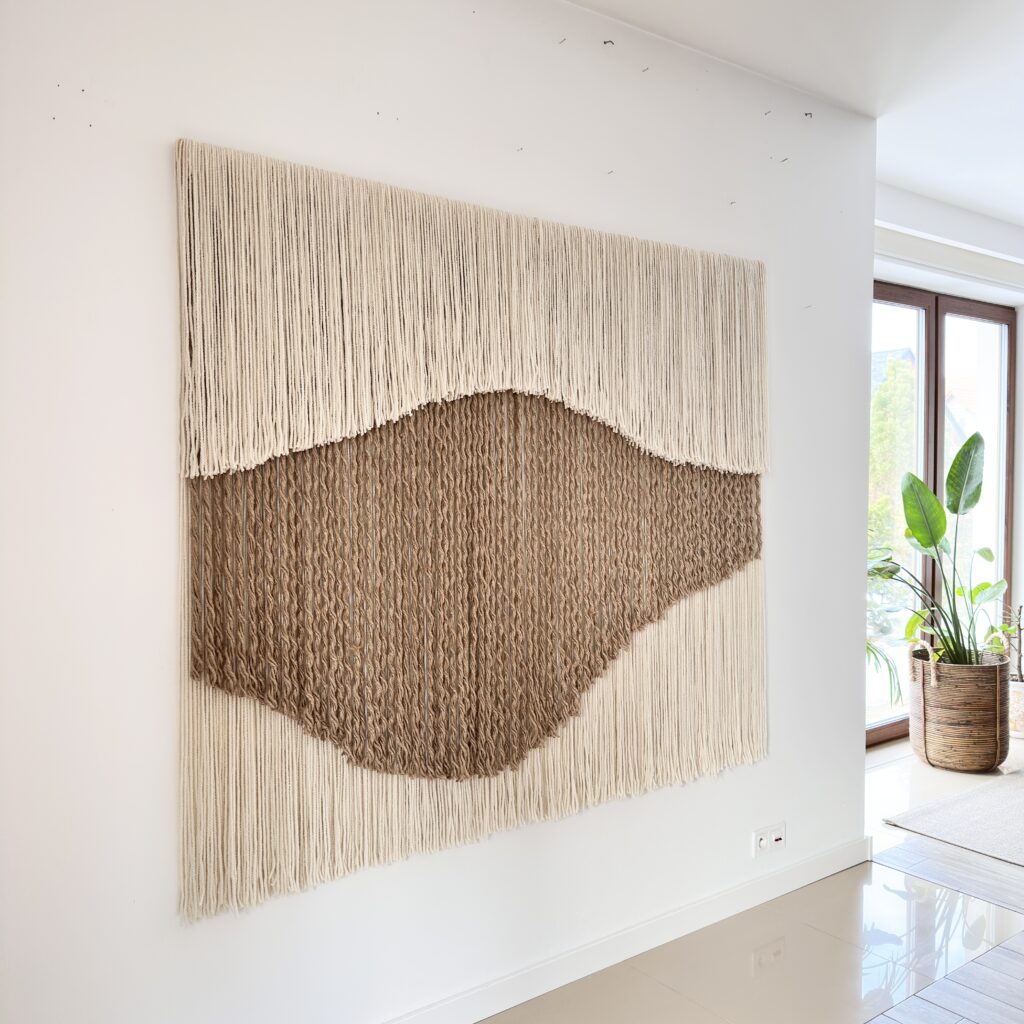 Large handwoven wool and jute wall hanging by Olivia Fiber Art displayed in a bright interior with a plant, highlighting natural textures and organic design.