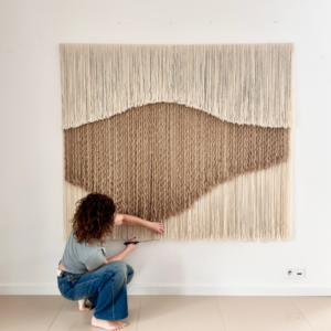 Artist installing a large handwoven wool and jute wall hanging by Olivia Fiber Art, showing the scale, craftsmanship, and one-of-a-kind nature of the textile artwork.