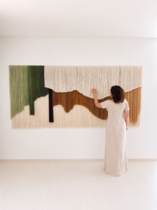 Large handwoven fiber art wall hanging made from hand-dyed alpaca and sheep wool displayed in a modern organic interior with woman standing beside the tapestry, luxury oversized textile wall decor in earthy neutral tones.